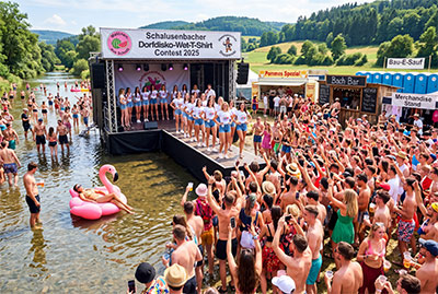 Contest am Flussufer. Auf einer Bühne stehen Frauen in weißen T-Shirts. Großes Publikum. Fressbuden.